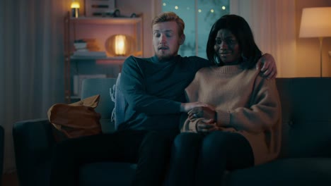 Happy-Young-Couple-Watching-Horror-Movie-on-TV-while-Sitting-on-a-Couch,-they-got-Scared,-Girl-Covers-eyes.-Handsome-Caucasian-Boy-and-Black-Girl-in-Love-Spending-Time-Together.-Moving-Camera-Shot.