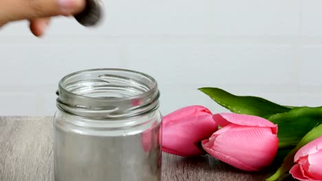 A-woman-is-taking-a-coin-in-a-jar,-which-is-placed-on-a-wooden-table-and-has-a-pink-tulip