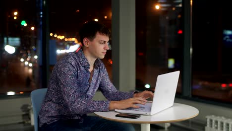 Un-hombre-escribiendo-en-un-teclado-de-ordenador-portátil,-en-la-oficina-en-una-gran-ventana,-contra-la-ciudad-de-noche.