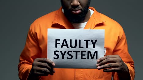 Faulty-system-phrase-on-cardboard-in-hands-of-Afro-American-prisoner,-disorder