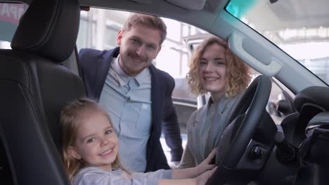 happy-childhood,-sweet-kid-girl-behind-wheel-of-automobile-together-with-mother-and-father-while-buying-family-machine-at-sales-center-close-up