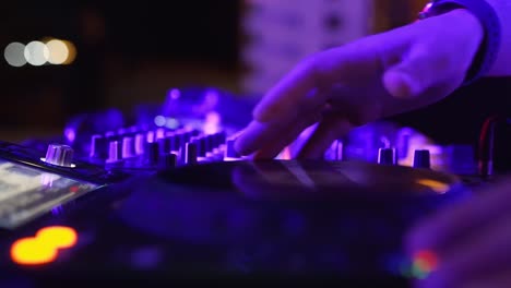 Las-manos-DJ-controlan-la-consola-de-sonido-para-mezclar-música-en-el-club-de-discoteca.-Botones-táctiles-de-los-dedos,-reproducción-de-música-electrónica-en-la-cubierta.-Las-luces-de-color-muestran-en-la-fiesta-de-baile-de-un-club-nocturno.-De-cerca-