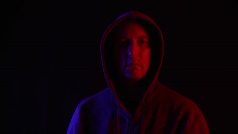 Mann-in-dunklem-Hoodie,-der-auf-schwarzem-Hintergrund-in-blauer-und-roter-Beleuchtung-zur-Kamera-aufschaut.-Porträt-mysteriösen-Mann-in-Kapuze-in-dunklen-Studio-mit-roter-und-blauer-Hintergrundbeleuchtung.