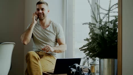 Attractive-young-man-talking-phone-sitting-on-windowsill-with-laptop-and-camera-at-home