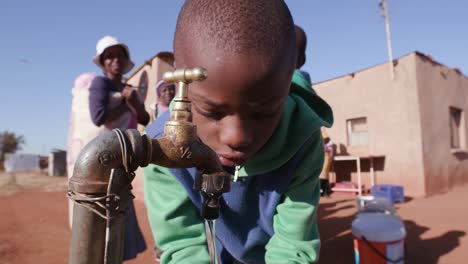 Primer-plano-del-joven-africano-bebiendo-agua-de-un-grifo-mientras-mujer-en-fila-para-recoger-agua-en-envases-de-plástico-debido-a-la-sequía-en-África-del-sur