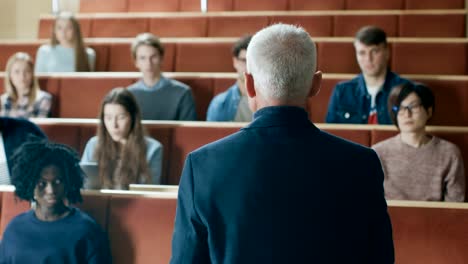 Clase-de-frente-a-cámara:-Prominente-profesor-escribiendo-en-la-pizarra-y-dar-Conferencia-a-un-aula-llena-de-estudiantes-Multi-étnica.-Universidad-moderna-con-jóvenes-brillantes-en-el-aula.