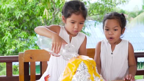 Two-asian-little-girls-do-the-baking-soda-and-vinegar-volcano-experiment-at-the-table-in-their-house,-slow-motion-in-50-fps