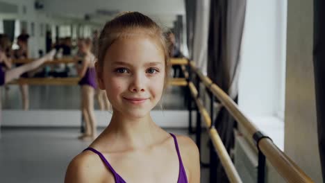 Close-up-portrait-of-beautiful-little-girl-in-bodysuit-standing-in-ballet-class,-smiling-and-looking-at-camera.-Other-students-are-doing-exercises-in-background.