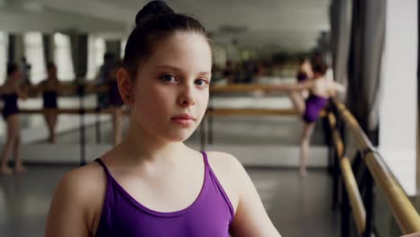 Primer-plano-retrato-de-niña-morena-en-traje-de-pie-en-clase-de-ballet-y-mirando-a-cámara.-Otros-estudiantes-están-practicando-en-fondo.