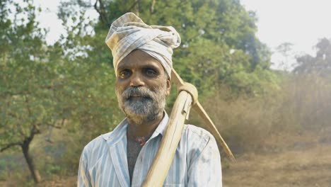 Un-hombre-con-barba-en-su-tierra-agrícola-rodeado-de-árboles-con-azada.