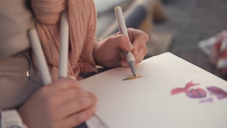 Mujer-es-dibujando-motivos-florales-en-un-sketchbook,-sentado-en-un-jardín,-primer-plano