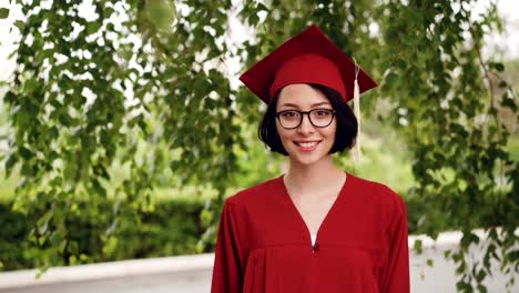 Porträt-der-fröhliche-Junge-Frau-Studium-Student-in-Robe-und-Doktorhut-lächelnd-und-Blick-auf-Kamera-stehend-unter-dem-Baum-auf-dem-Campus.-Jugend-und-Bildung-Konzept.