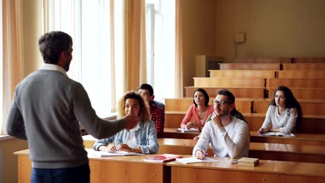 Grupo-multiétnico-de-jóvenes-es-escuchar-al-maestro-y-sonriendo-levantando-manos,-profesor-es-apuntando-a-la-chica-y-hablar-con-ella.-Concepto-de-alumnos-y-tutores.