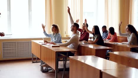 Friendly-teacher-is-talking-to-students-asking-questions-while-young-people-are-raising-hands-and-answering-sitting-at-desks.-High-school,-education-and-youth-concept.