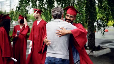 Stolz-auf-Lehrer-ist-Student,-seine-Hand-zu-schütteln-und-umarmt-ihn-beim-Abschluss-Tag-stehen-auf-dem-Campus-in-traditionellen-Grad-Kleidungsstück-gratulieren.-Bildung-und-Menschen-Konzept.