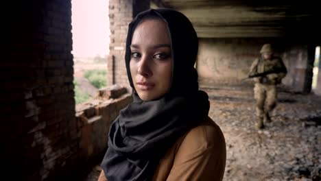 Young-muslim-woman-in-hijab-crying-and-looking-at-camera,-soldier-with-gun-walking-in-background,-abandoned-building,-terrorism-concept