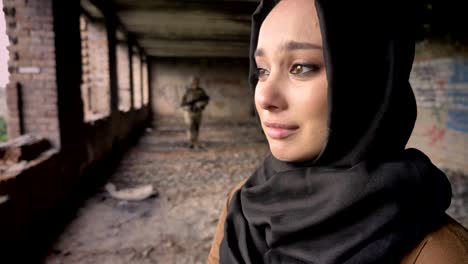 Young-sad-muslim-woman-in-hijab-crying-when-armed-soldier-going-towards-woman,-abandoned-building,-war-concept