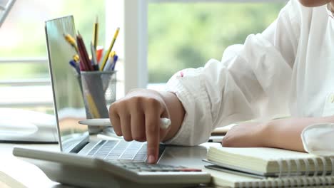 Student-learning-online-study-concept:-Beautiful-Asian-girl-learning-and-writing-in-textbook-or-homework,-short-notes-with-laptop,-pencils-box-on-desk-in-home-for-e-learning-near-window-background