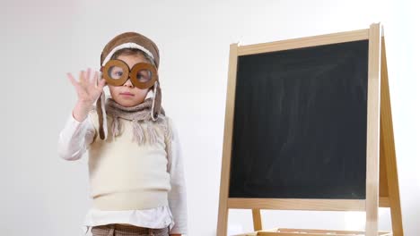 Una-pequeña-niña-vestida-como-un-aviador-o-piloto,-indica-con-su-mano-la-pizarra-detrás-de-ella-como-un-iniciar-vuelo-para-aprender-a-usar-el-avión-y-la-imaginación.