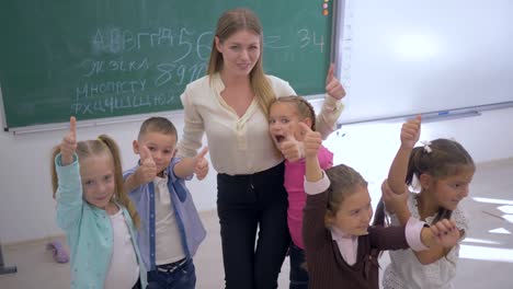 happy-schoolchilds-with-teacher-showing-thumbs-up-in-classroom-on-background-of-board-at-school