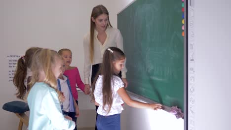 pupils-and-teacher-standing-near-whiteboard-in-classroom,-schoolgirl-takes-piece-of-chalk-and-writes-answer-at-the-lesson