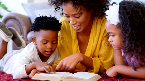 Vista-frontal-de-negro-de-la-madre-con-sus-hijos-leyendo-un-libro-en-un-cómodo-hogar-4k