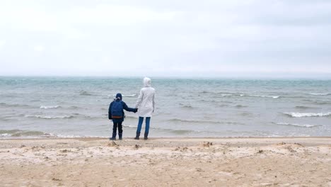 Mom-and-son-are-walking-on-the-sea-sand-beach-in-winter,-back-view.