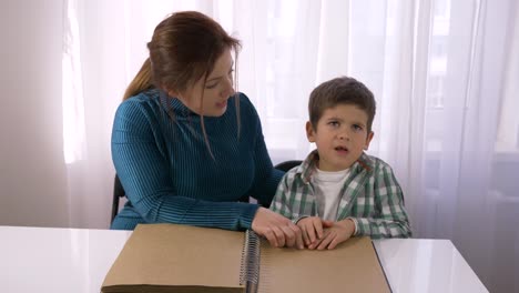 difficult-schooling-for-blind-children,-young-teacher-teaches-Visually-impaired-boy-to-read-braille-books-with-symbols-font-sitting-at-table-in-room