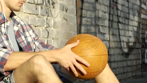 Young-man-listening-to-music-and-spinning-basket-ball-in-hands,-waiting-friends
