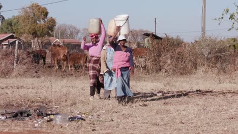 Three-african-woman-carrying-water-on-their-heads-in-plastic-buckets-from-a-remote-source-and-walking-back-to-their-homes