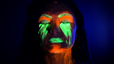 Porträt-eines-Mädchens-schmiert-und-fluoreszierende-Farbe-unter-den-Augen-reibt.-Farbstoff-in-der-Nähe-von-UV-Schwarzlicht-Leuchten.-Frau-mit-Zöpfen-im-Neonlicht.-Night-Club,-Party,-Halloween-psychedelischen-Konzepte