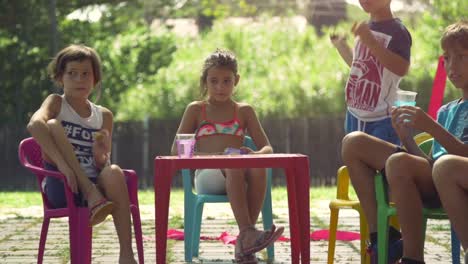 Young-happy-kids-in-summer-camp-sitting-colorful-plastic-chairs-drinking-eating-snacks-having-fun-outside-in-green-grass-field-children-party-slow-motion