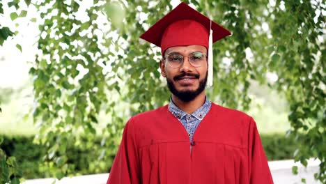 Portrait-of-bearded-mixed-race-man-graduating-student-in-gown-and-mortar-board-smiling-and-looking-at-camera-standing-outdoors-on-campus.-People-and-education-concept.