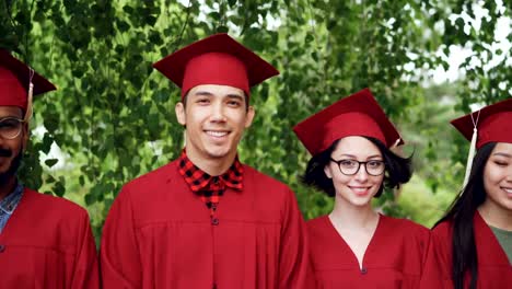 Portrait-of-multinational-group-of-graduating-students-in-red-graduation-gowns-and-mortar-boards-standing-together-outdoors,-smiling-and-looking-at-camera.