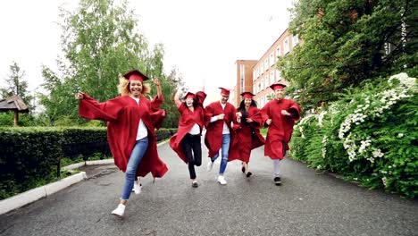 Dolly-Schuss-von-aufgeregt-Absolventen-auf-dem-Campus-tragen-Kleider-und-Trachtenhüte-feiert-Ende-des-Studiums.-Höhere-Bildung,-Jugend-und-Glück-Konzept.