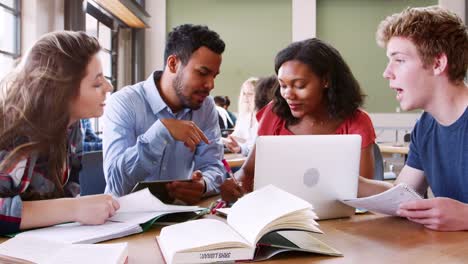 SchülerInnen-und-Schüler-mit-Laptops-und-digitalen-Brettchen-mit-männlichen-Lehrer-am-Schreibtisch-arbeiten