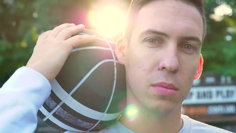 Portrait-of-young-confident-man-holding-basketball-and-looking-at-camera,-park-in-background