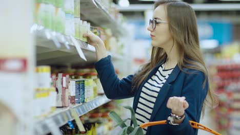 Im-Supermarkt:-schöne-junge-Frau-blättert-durch-die-Konserven-Abschnitt-des-Speichers.-Sie-hat-Warenkorb-voll-von-gesunden-Lebensmitteln.