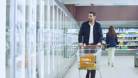 En-el-supermercado:-guapo-empuja-compras-la-tarjeta-y-navega-por-los-productos-en-la-sección-de-congelados.-Hombre-busca-en-refrigerador-de-puerta-de-cristal,-elegir-productos-lácteos.-Otro-cliente-comercial.