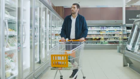 En-el-supermercado:-guapo-empuja-compras-la-tarjeta-y-navega-por-los-productos-en-la-sección-de-congelados.-Otro-cliente-en-el-fondo.