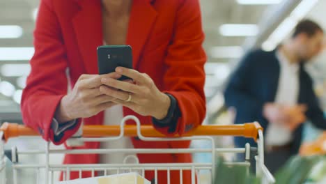 Im-Supermarkt:-Frau-nutzt-Smartphone,-lehnt-sich-auf-Ihren-Warenkorb.-In-der-großen-Mall-Frau-Surfen-im-Internet-auf-ihr-Handy.-Hände-halten-Handys-im-Mittelpunkt.