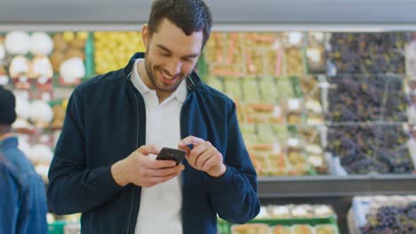 At-the-Supermarket:-Handsome-Man-Uses-Smartphone-while-Standing-in-the-Fresh-Produce-Section-of-the-Store.-Man-Immersed-in-Internet-Surfing-on-His-Mobile-Phone-In-the-Background-Colorful-Fruits-and-Organic-Vegetables.