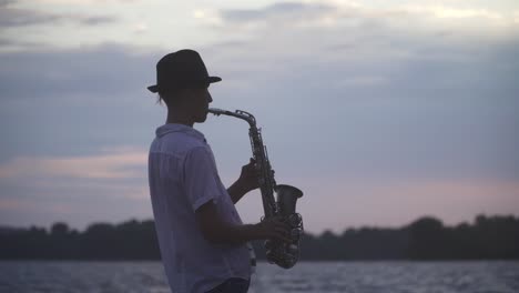 Chico-joven-guapo-es-jugar-el-saxophone-en-la-orilla-del-río