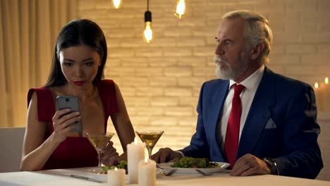 Girl-sitting-in-restaurant-with-boring-old-man,-chatting-on-phone,-bad-date