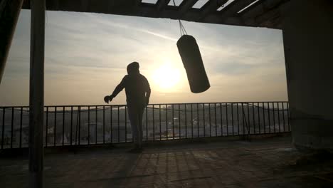 Boxing-bag-swinging,-man-comes-on-terrace-and-looks-at-city,-sunset,-silhouettes,-sportsman-practicing,-power-training,-strong-guy-hard-exercising,-strength-exercises,-workout,-handheld,-sunny-day.