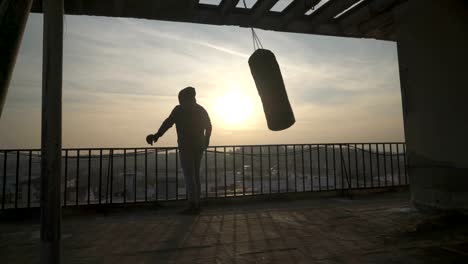 Box-Tasche-geschwungen,-Mann-kommt-auf-Terrasse-und-schaut-auf-Stadt,-Sonnenuntergang,-Silhouetten,-Sportler-üben,-Kraft-training,-starker-Kerl-hart-trainieren,-Kraft-Übungen,-Training,-handheld,-sonnigen-Tag.