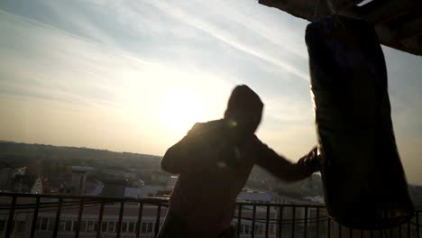 Boxer-performs-series-of-punching-in-boxing-bag-during-power-training-in-sunset,-sportsman-practicing,-power-training,-strong-guy-hard-exercising,-strength-exercises,-workout,-handheld,-sunny-day.