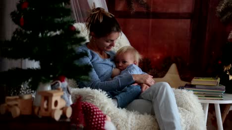 Mother-breastfeeding-her-toddler-son-sitting-in-cozy-armchair-near-Christmas-tree,-wintertime