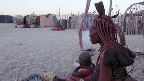 4K-side-view-of-Himba-woman-in-traditional-dress-with-young-child,-sitting-and-smoking-outside-their-hut-within-their-small-compound,-Namibia