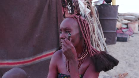 4K-close-up-view-of-Himba-woman-in-traditional-dress-with-young-child,-smoking-a-pipe-outside-their-hut-within-their-small-compound,-Namibia